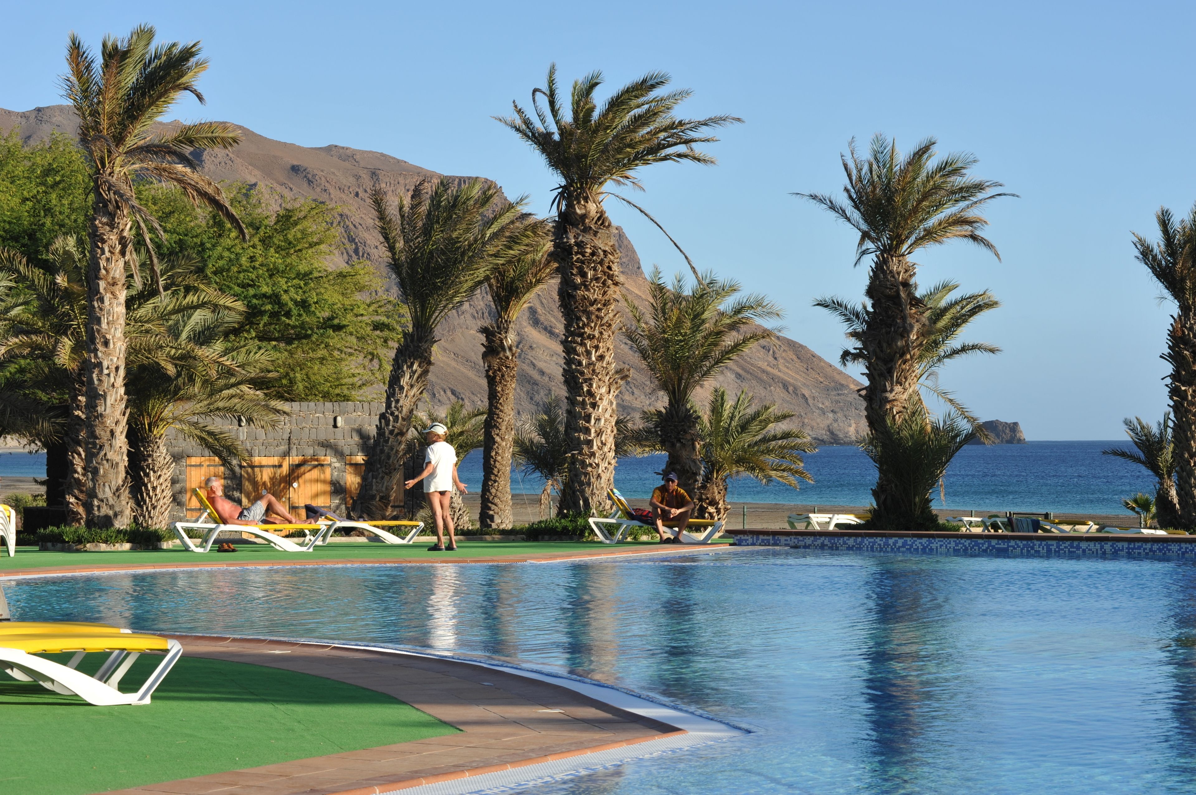 Ein Poolbereich mit Palmen, Liegestühlen und Blick auf das Meer und die Berge.