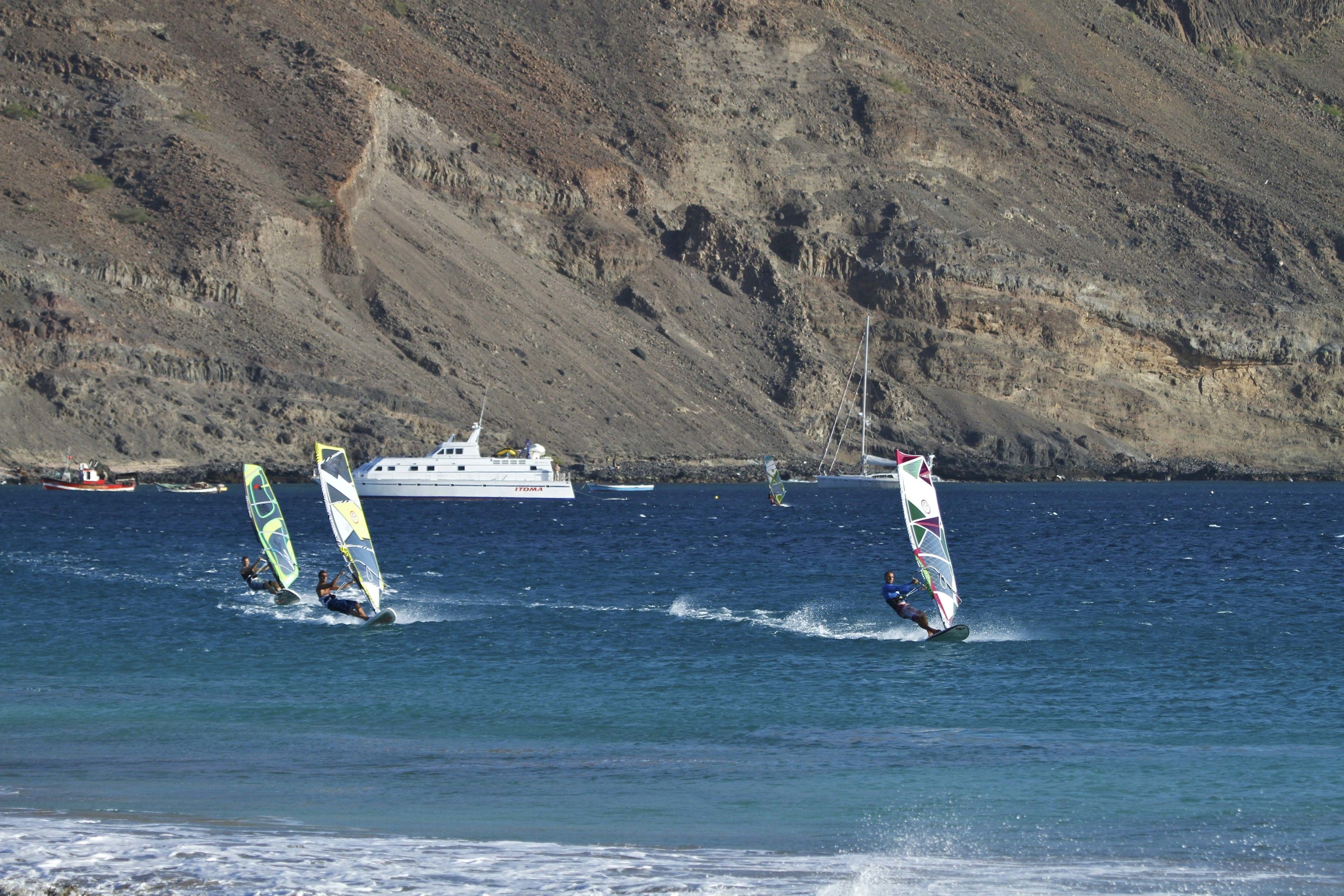 Paisaje costero con windsurfistas en el agua, un yate al fondo y una ladera rocosa.