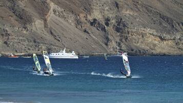 Paisaje costero con windsurfistas en el agua, un yate al fondo y una ladera rocosa.