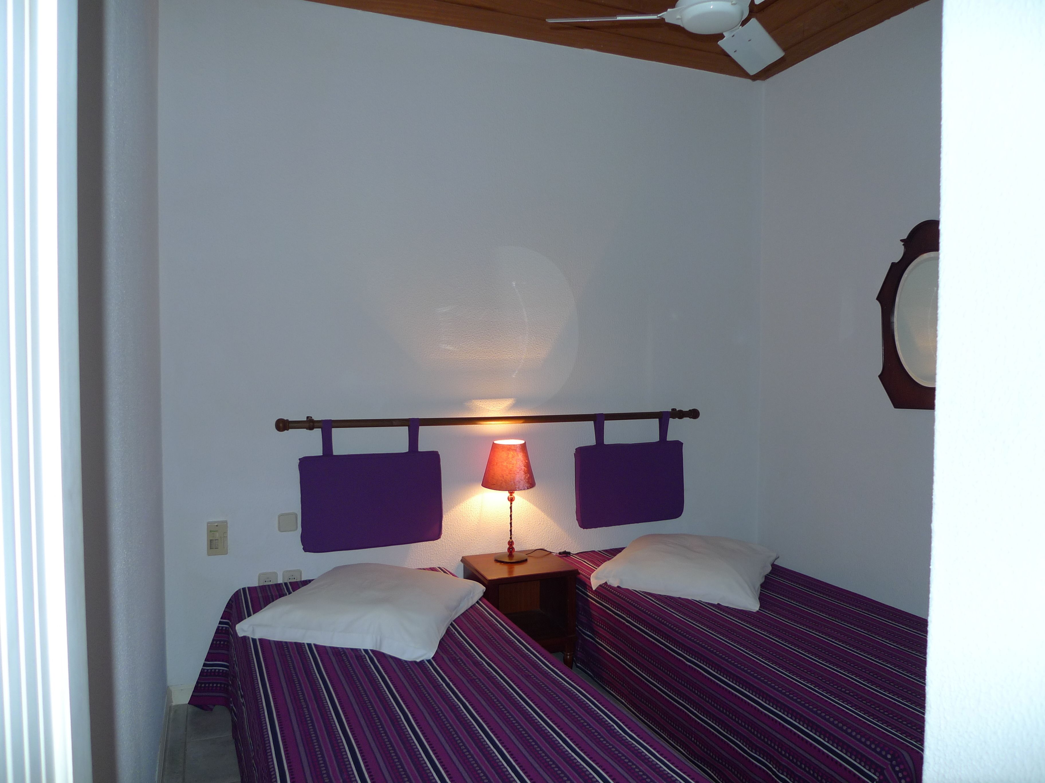 A small, single-room hotel room with two beds, a nightstand, a lamp, and a ceiling fan.