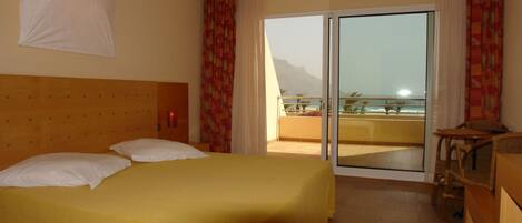 A hotel room with a bed, a balcony with a view of the sea and mountains, and a wooden headboard.