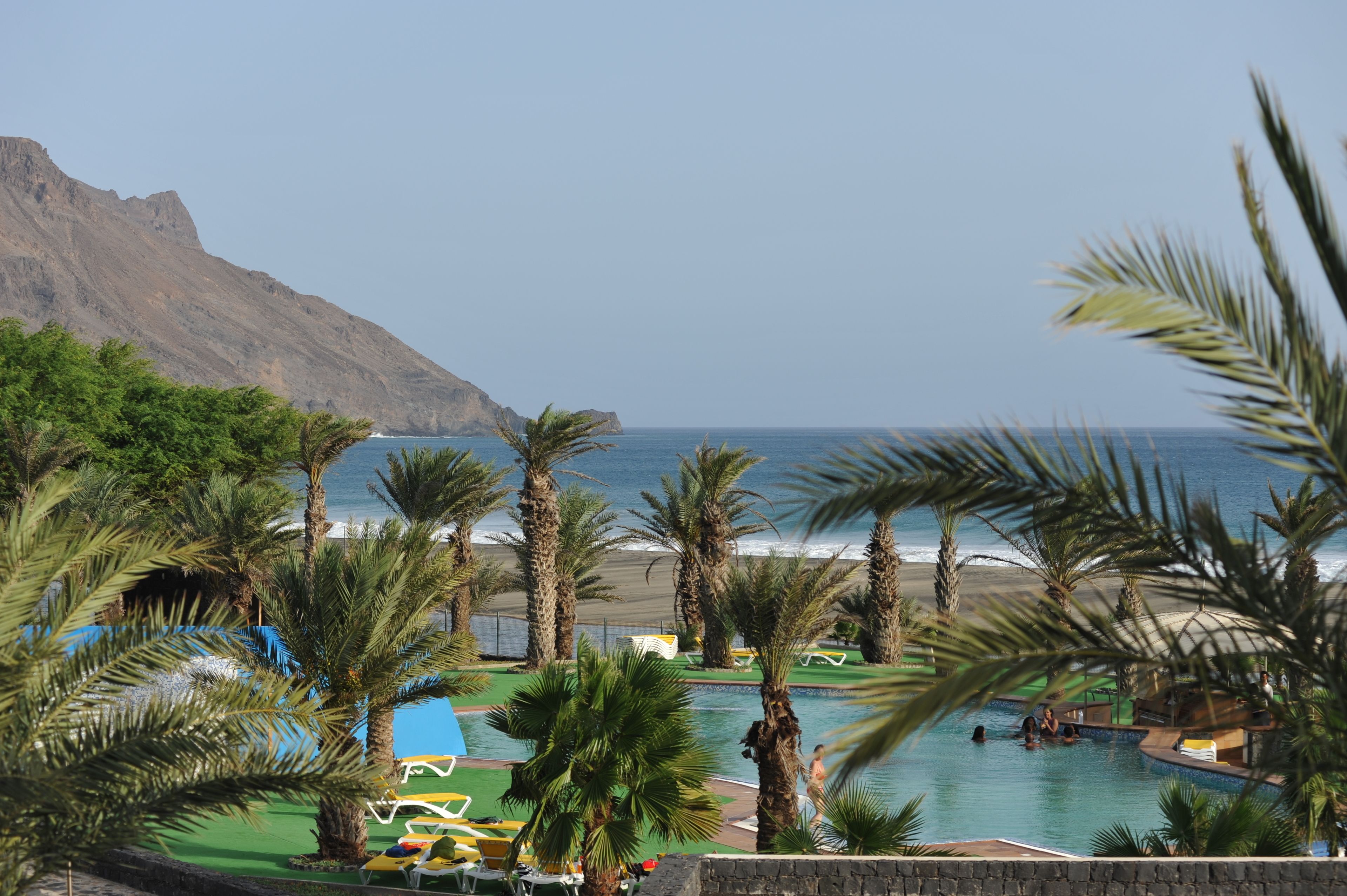 A pool area with palm trees, a beach, and a mountain in the background.