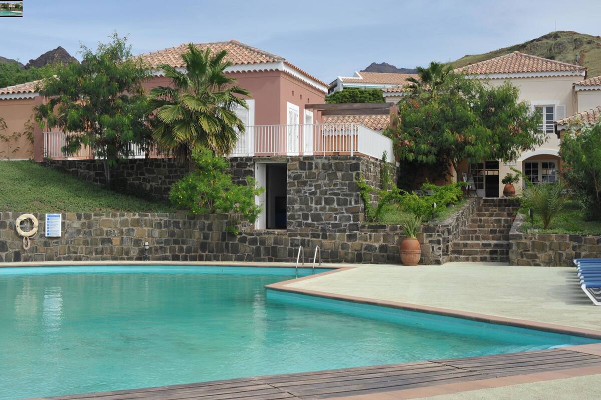 A swimming pool with a wooden deck, stone walls, and a view of houses with tiled roofs and palm trees.