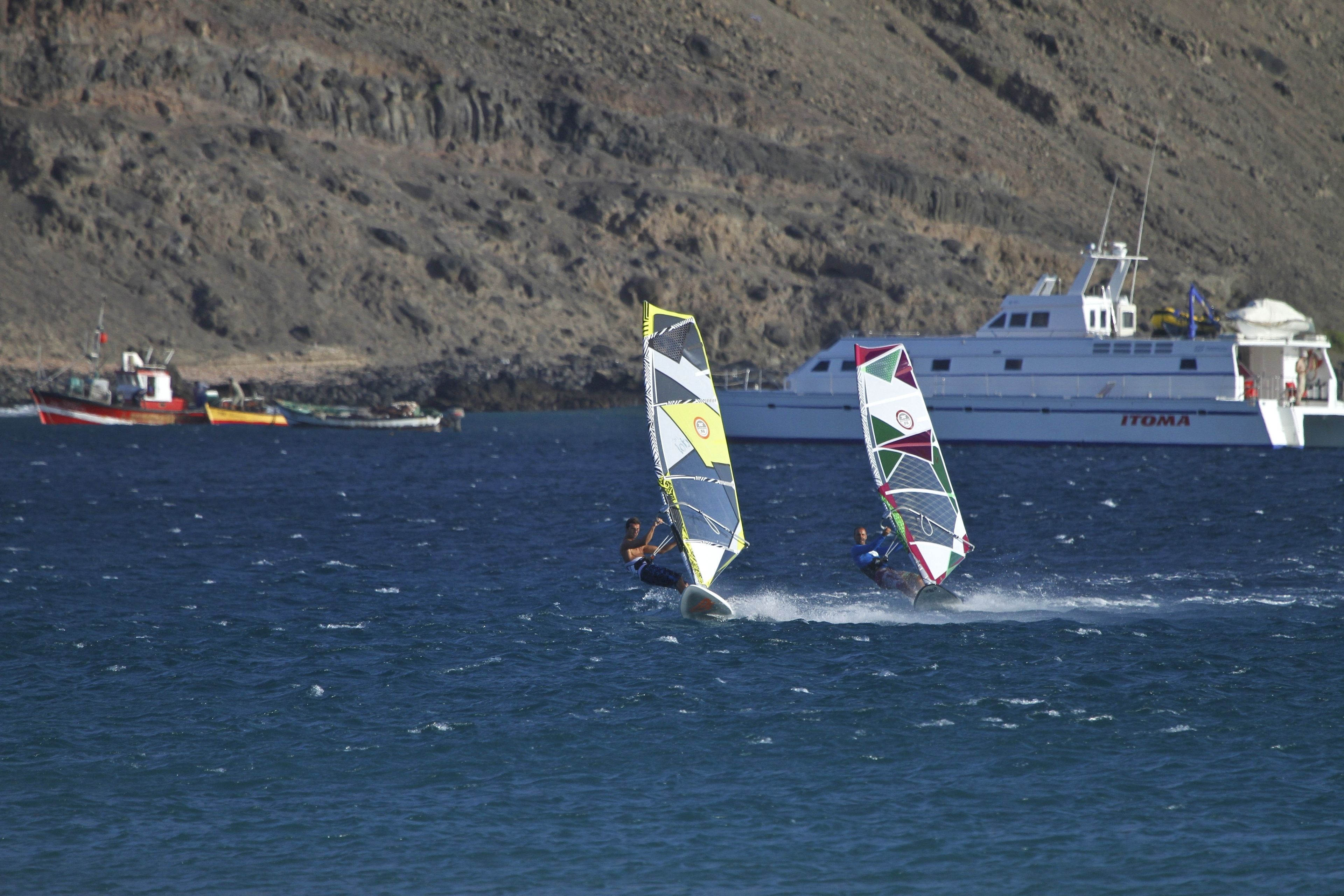 Two windsurfers on the water with a boat and rocky hills in the background.