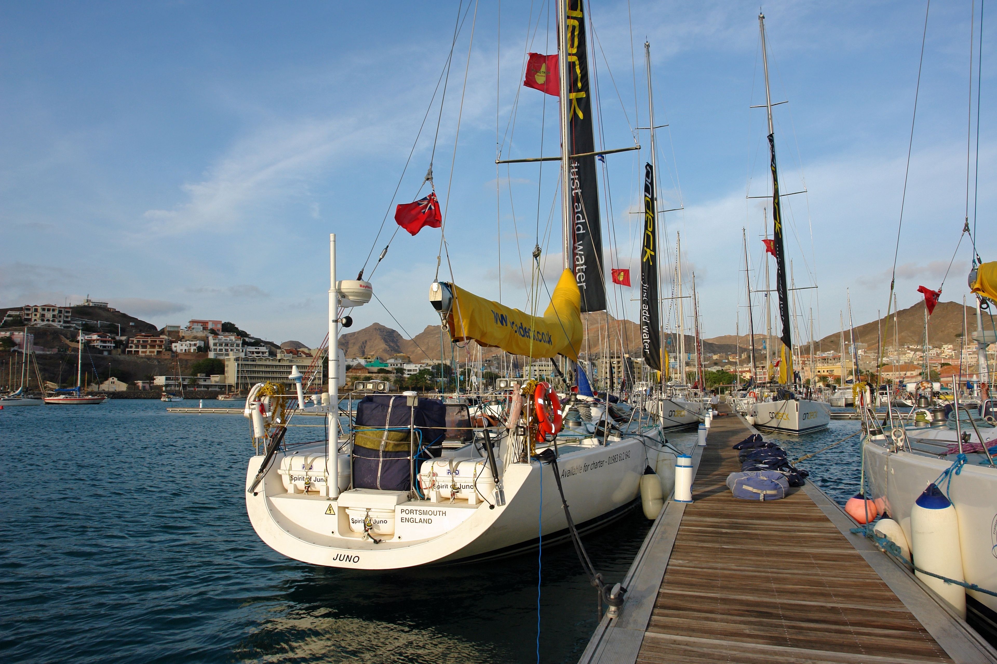 Un port de plaisance où sont amarrés plusieurs voiliers, une jetée en bois et, en arrière-plan, une vue sur la ville.