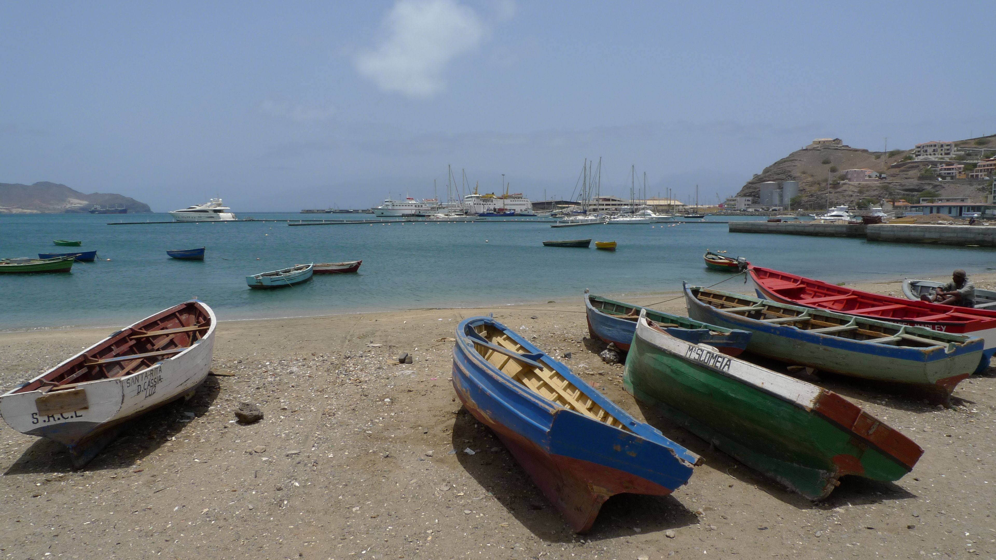 A beach with several small boats, a sandy shore, and a distant view of larger boats and buildings.