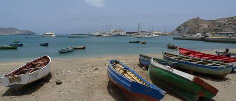A beach with several small boats, a sandy shore, and a distant view of larger boats and buildings.
