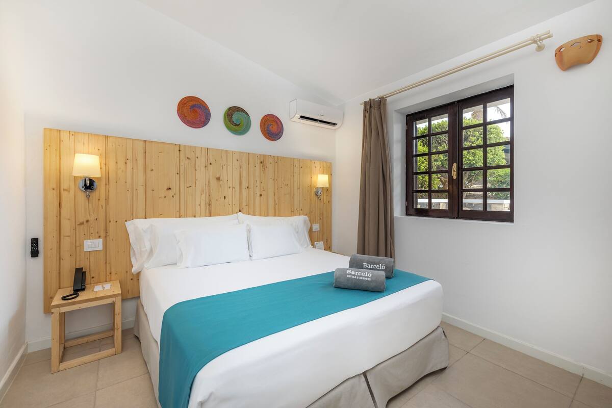 Habitación de hotel con cabecera de madera, cama con colcha color turquesa, ventana con vista a follaje y aire acondicionado de pared.