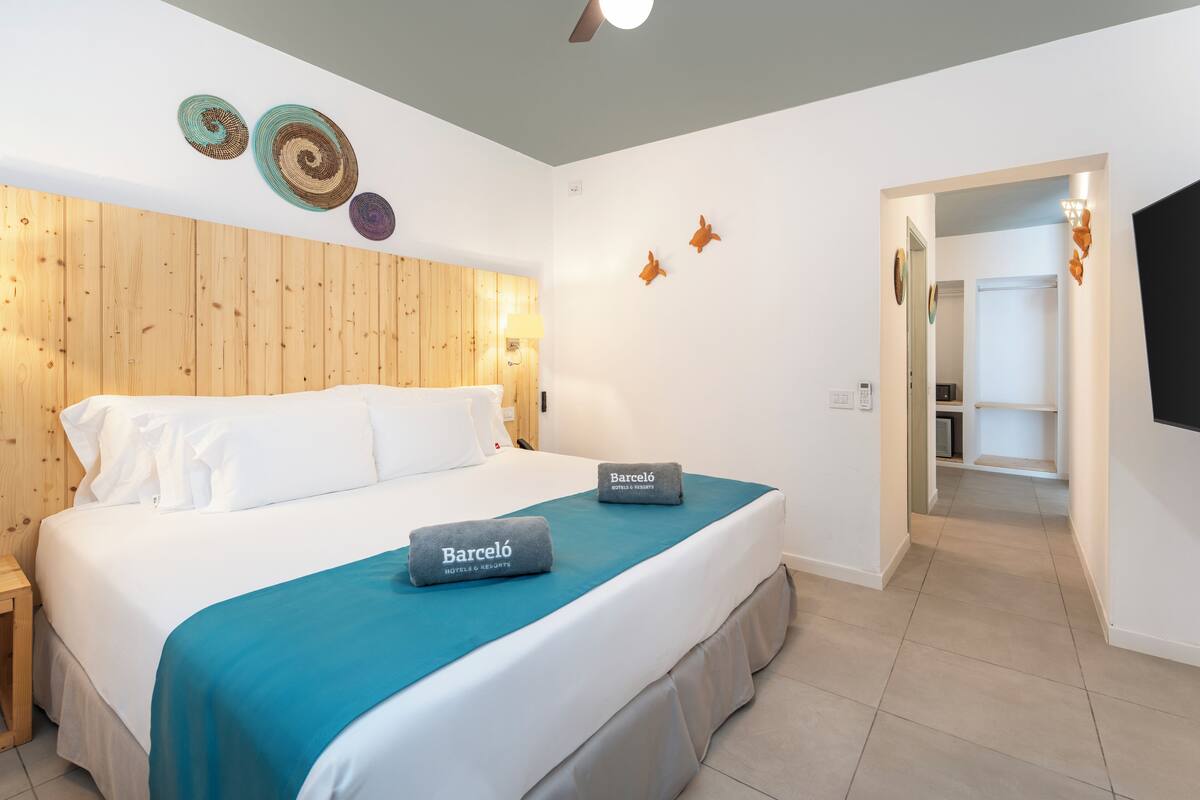 Habitación de hotel con cabecera de madera, cama con colcha color turquesa y dos almohadas con el nombre del hotel. Se observan placas decorativas en la pared y una puerta que conduce a otra habitación.