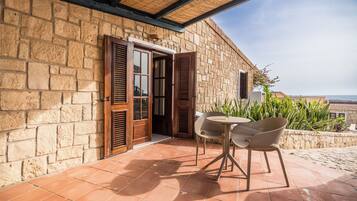 A stone building with a wooden door and shutters, a tiled patio with two chairs, and a view of the sea.