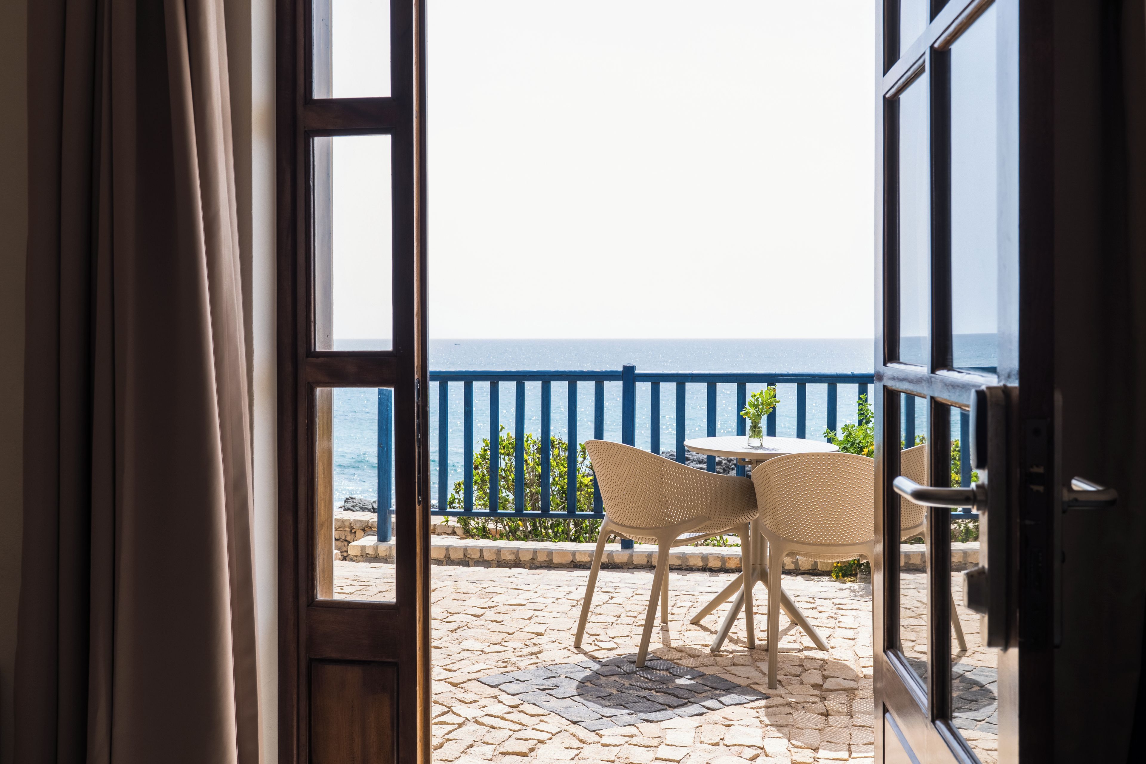 Eine offene Tür führt zu einem Balkon mit Tisch und Stühlen, der auf eine blaue Brüstung und einen Meerblick blickt.