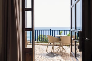 Une porte ouverte donnant sur un balcon équipé d’une table et de chaises, d’où l’on aperçoit une rambarde bleue et une vue sur la mer.