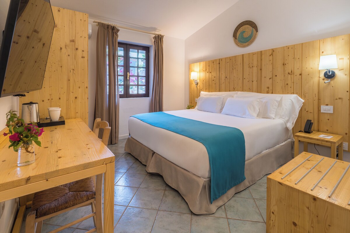 Een hotelkamer in houtstijl, met een bed met wit en blauw beddengoed, een houten bureau met een bloevas en een bijzettafeltje met een lamp.