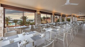 Un vaste espace extérieur aménagé pour les repas, avec des tables et des chaises, de grandes fenêtres offrant une vue sur les palmiers et la plage, et un ventilateur de plafond.