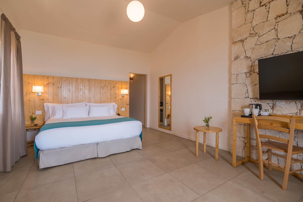 Ein Hotelzimmer mit einem Bett, Nachttischlampen, einem kleinen Tisch mit Stuhl, einem Spiegel und einem Flachbildfernseher, der an einer Steinwand befestigt ist.