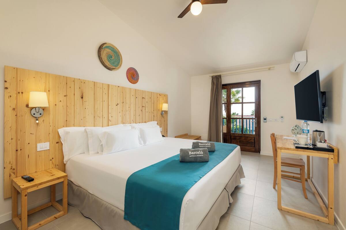 Habitación de hotel con cabecera de madera, cama con colcha color turquesa, dos mesitas de noche con lámparas, escritorio con silla, televisor y ventana con cortinas.