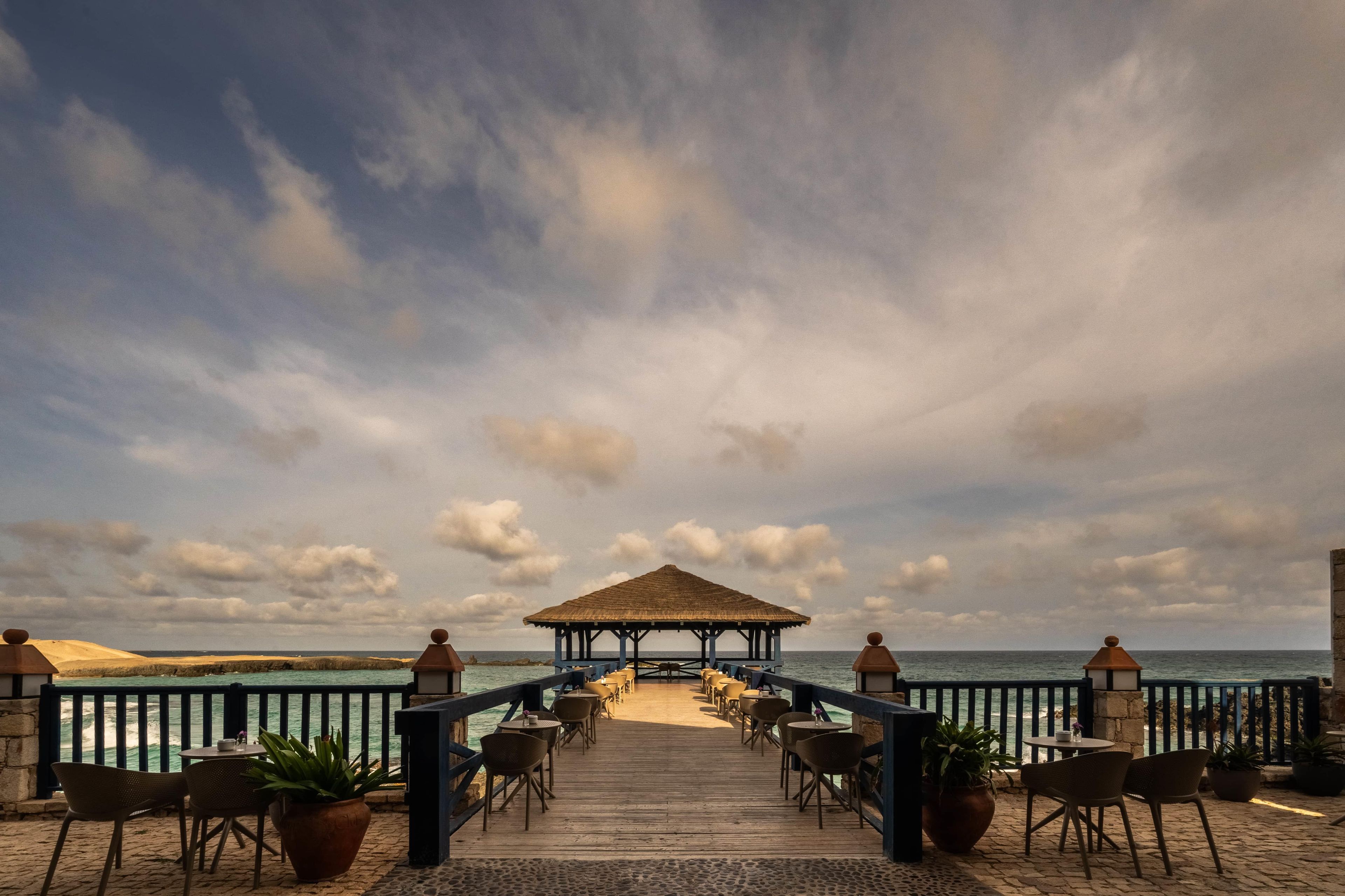 Une terrasse en bois mène à un pavillon à toit de chaume, doté de tables et de chaises, et offrant une vue sur la plage.