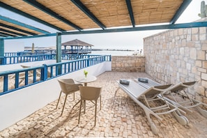Um pátio externo com mesa e cadeiras, uma mesinha com um vaso, e vista para o mar.