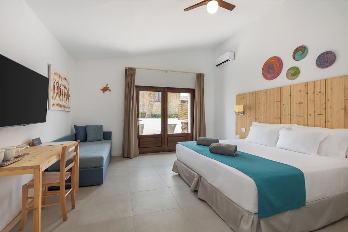 Una habitación de hotel con una cama, un escritorio con una silla, un sofá azul, un cabecero de madera y vistas a través de un ventanal.