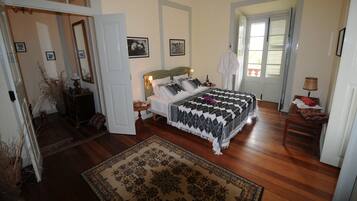 Un dormitorio con una cama grande, una mesita de noche, persianas en una ventana y una puerta que da a otra habitación.