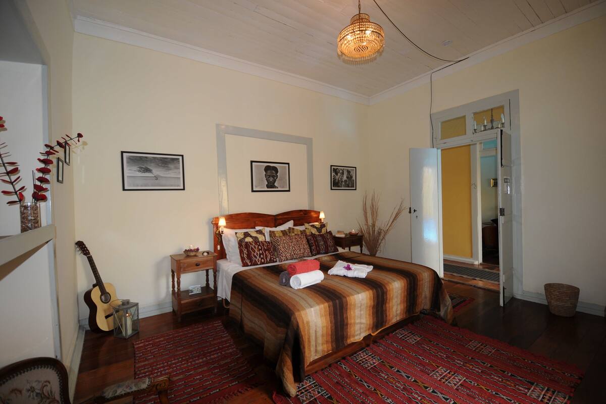 A bedroom with a large bed, a guitar, a mirror, and framed pictures on the wall.