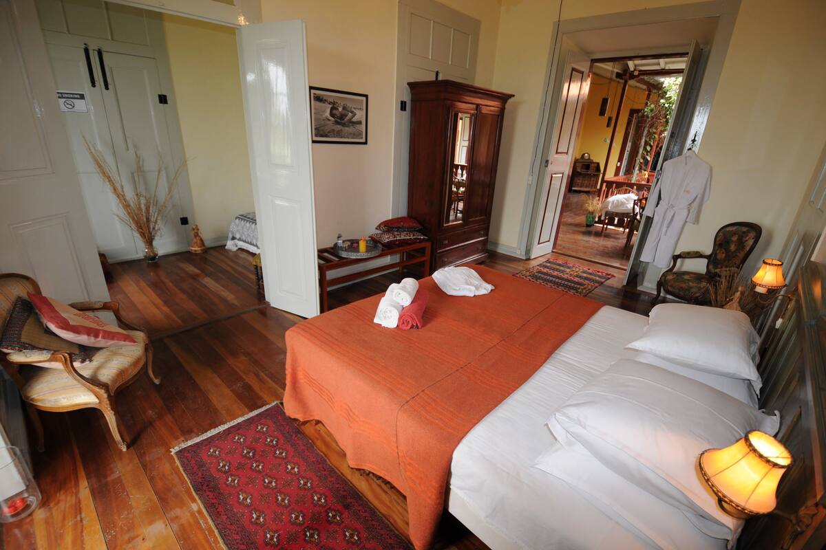 A hotel room with a large bed, a wooden wardrobe, a sitting area with a chair, and a small table with a vase and a tray.