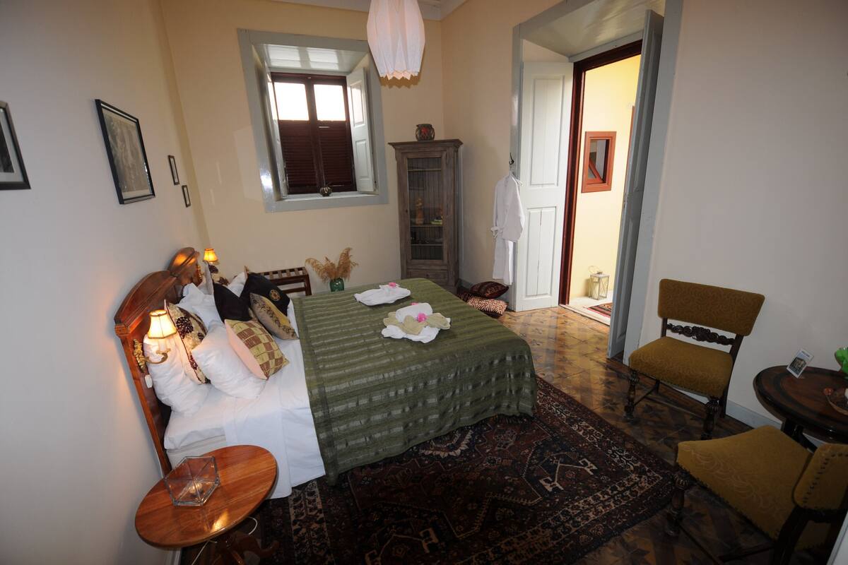 A bedroom with a bed, a nightstand, a chair, a window, a cabinet, and a door leading to another room.