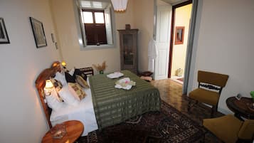 Un dormitorio con cama, mesita de noche, silla, ventana, armario y puerta que da a otra habitación.