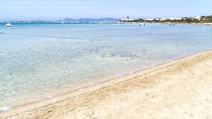 A beach with clear shallow water, a sandy shore, and distant buildings. - El Lago Dorado (Formentera)