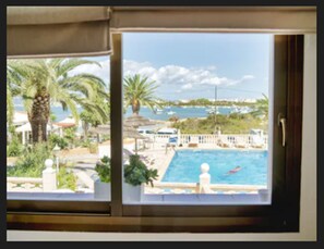 A view from a window overlooking a swimming pool with a clear sky and distant coastline. - El Lago Dorado (Formentera)