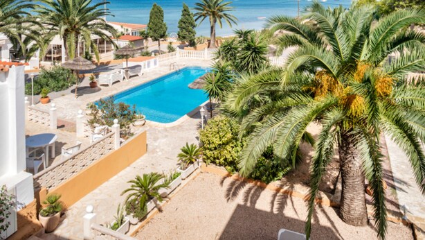 A pool area with palm trees, a clear blue pool, and a view of the sea and distant buildings. - El Lago Dorado (Formentera)