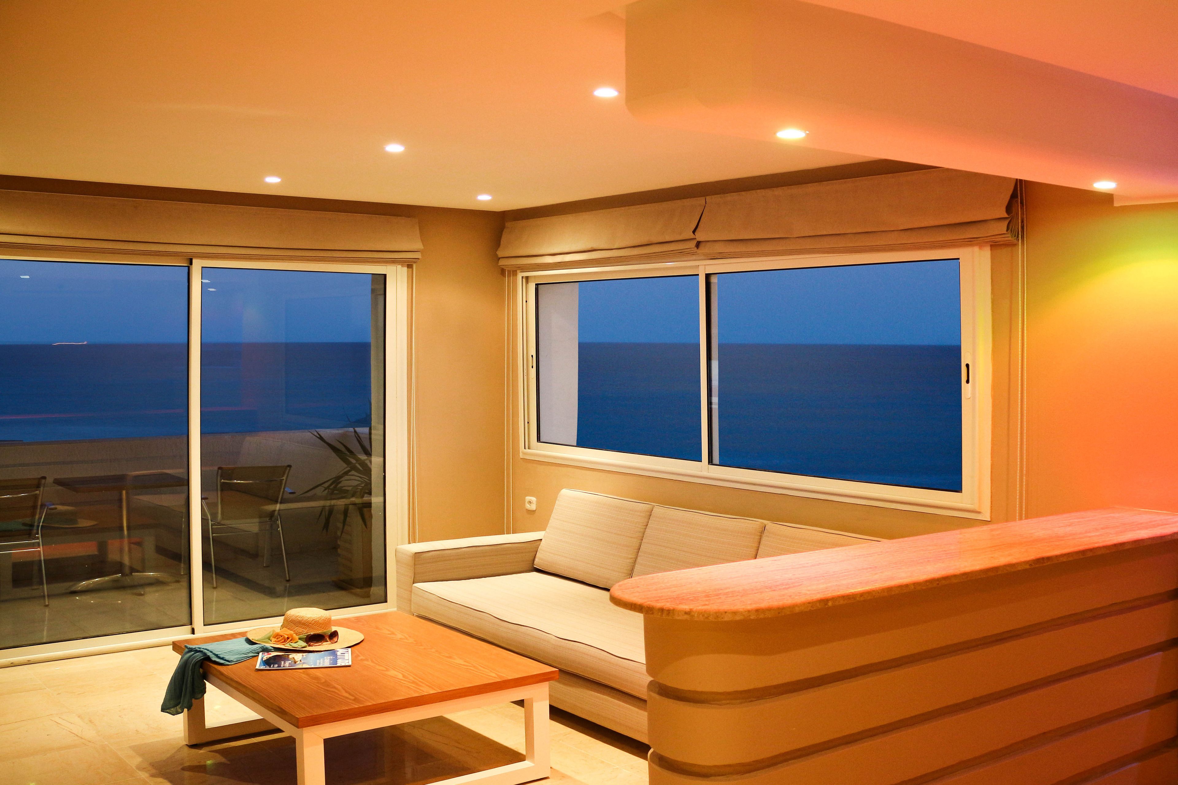 Een balkon met een bank en een tafel, uitzicht op zee, en een groot raam met gordijnen.