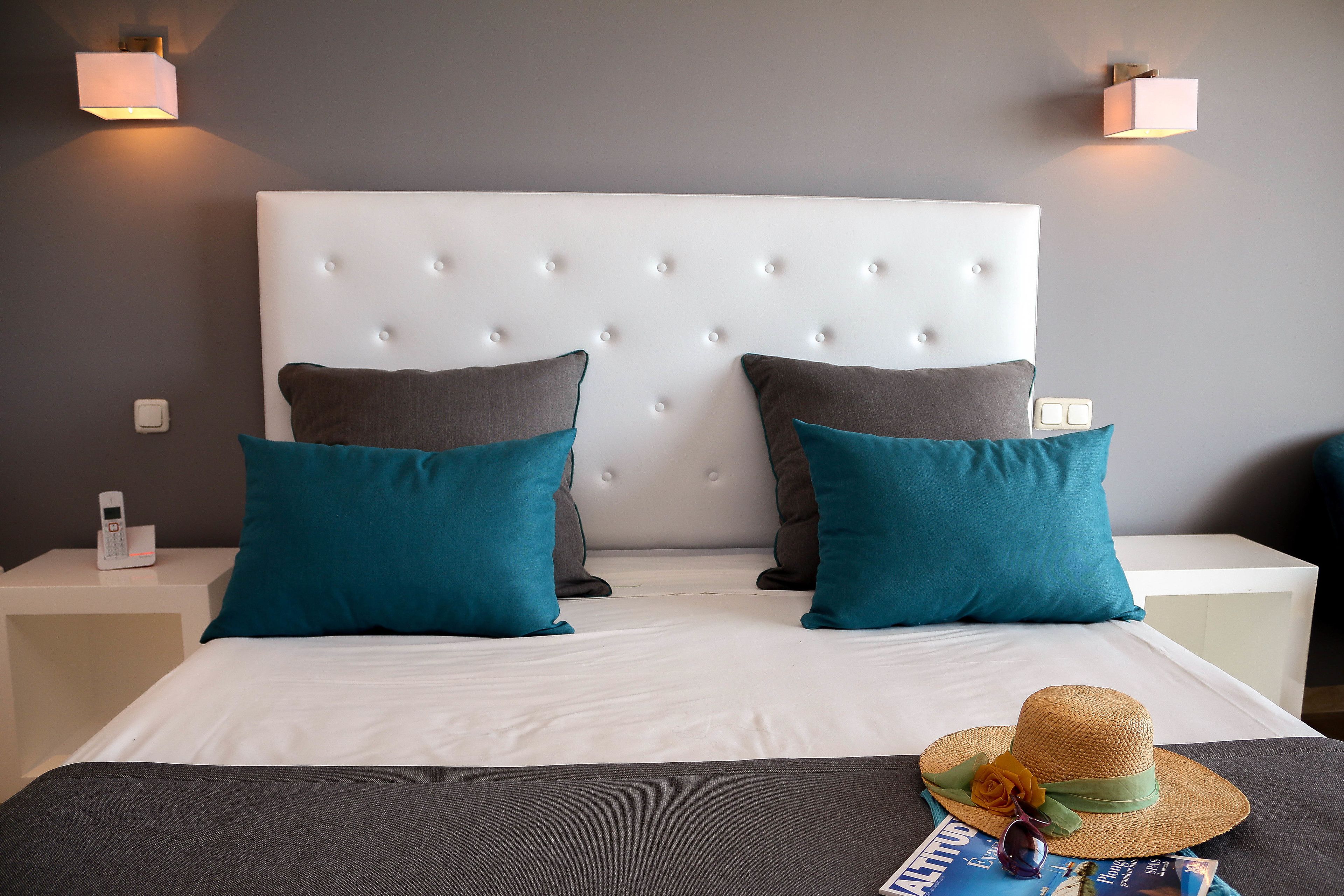 A neatly made bed with a white headboard, teal and gray pillows, and a straw hat on the bed.