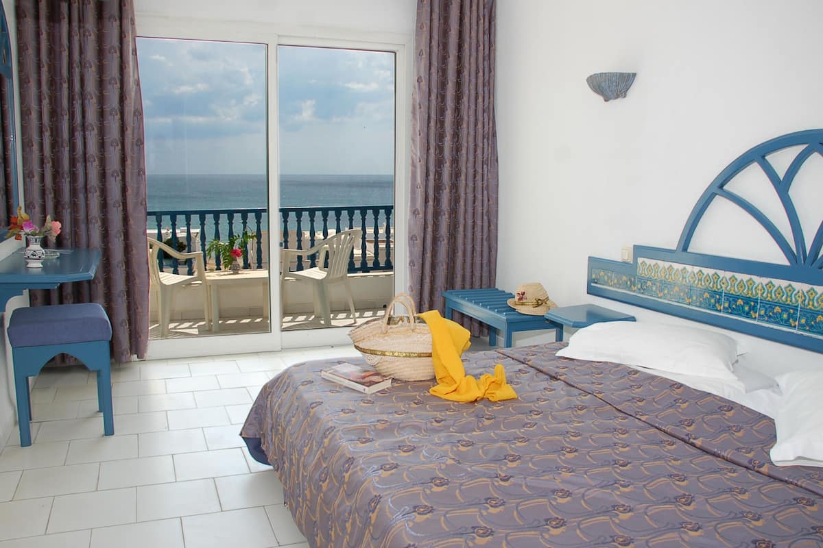 A bedroom with a bed, a desk, a chair, and a view of the sea through a glass door.