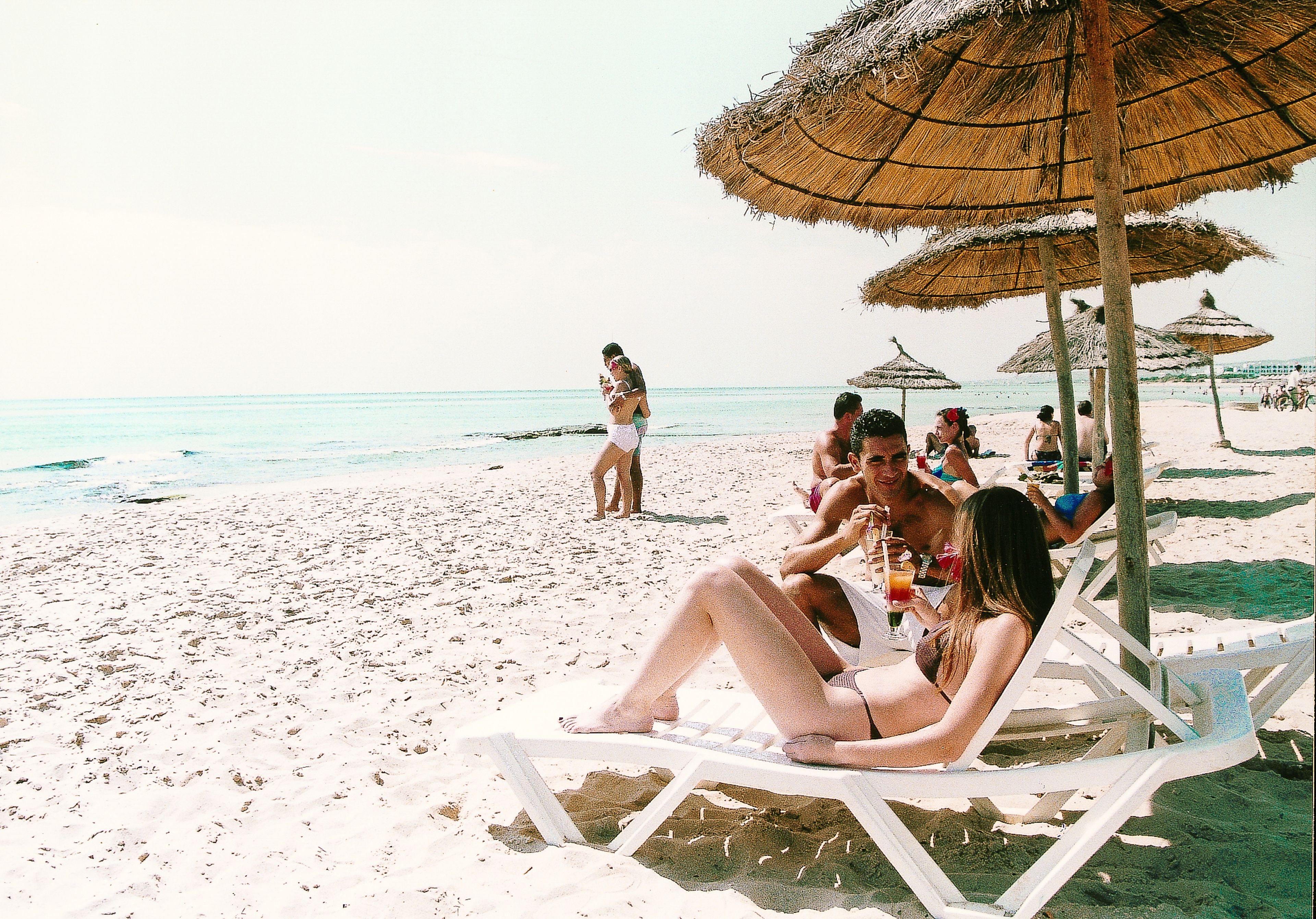 A beach scene with people relaxing under thatched umbrellas, some sitting on lounge chairs, and others walking along the shore.