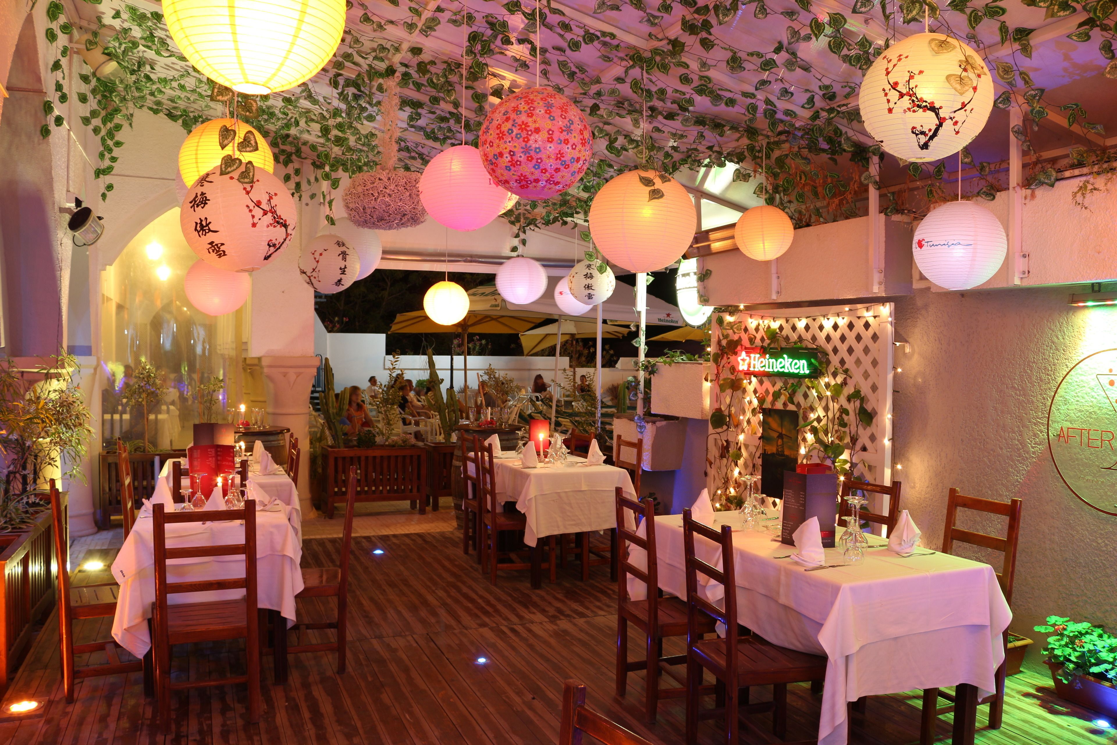 An indoor dining area with tables set for guests, hanging paper lanterns, and a Heineken sign.