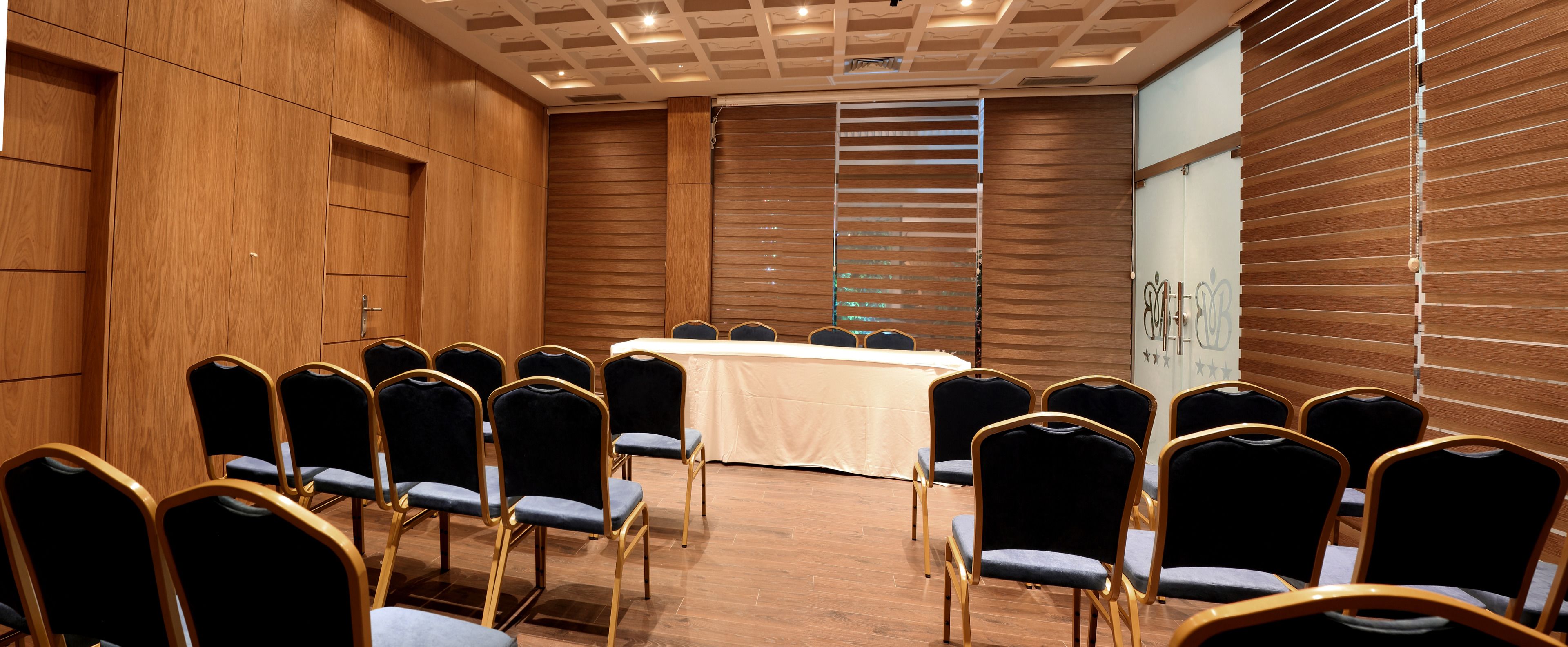 A conference room with a long table, rows of chairs, wooden paneling, and a glass partition with a logo.