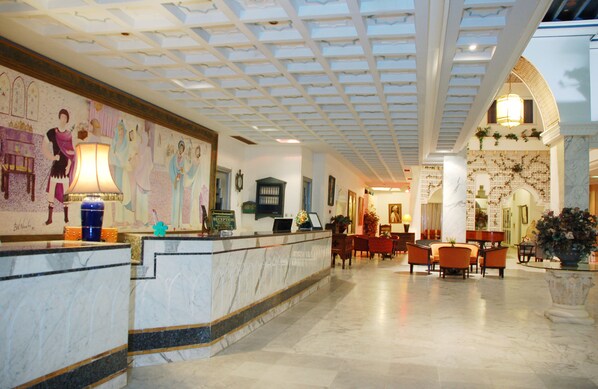 A hotel lobby with a reception desk, seating area, and decorative elements like a mural and plants.