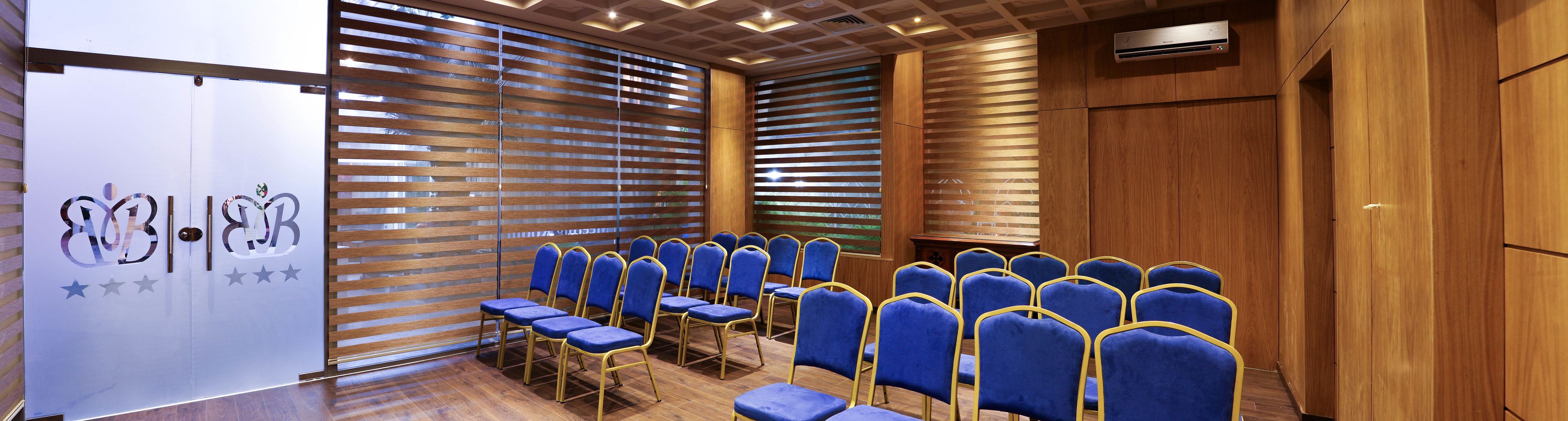Un salón de conferencias con hileras de sillas azules, paneles de madera y una pizarra con un logotipo.