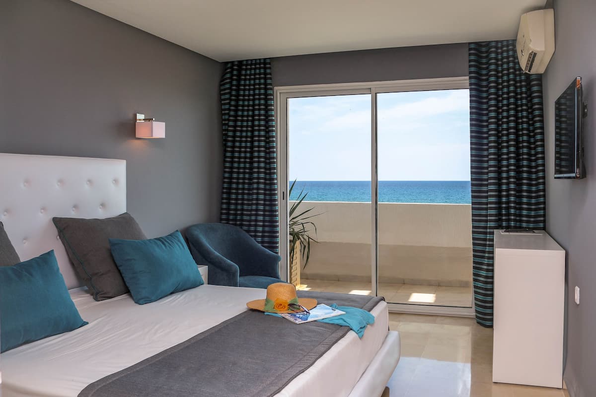 A bedroom with a large bed, a bedside table, a TV, and a view of the sea through a sliding door.