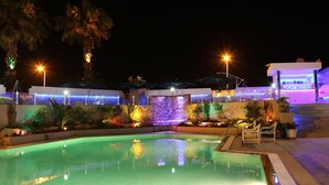 A nighttime pool area with a clear pool, surrounding plants, and a bar area with umbrellas. - Hotel Byzance (Nabeul)