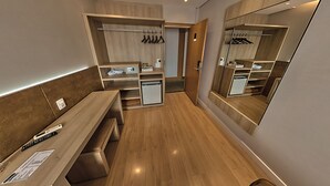 A modern hotel room with a wooden floor, a desk with a remote control, a mirror, and a built-in wardrobe with shelves and a hanging rack. - Shelton Hotel (Serra Negra)