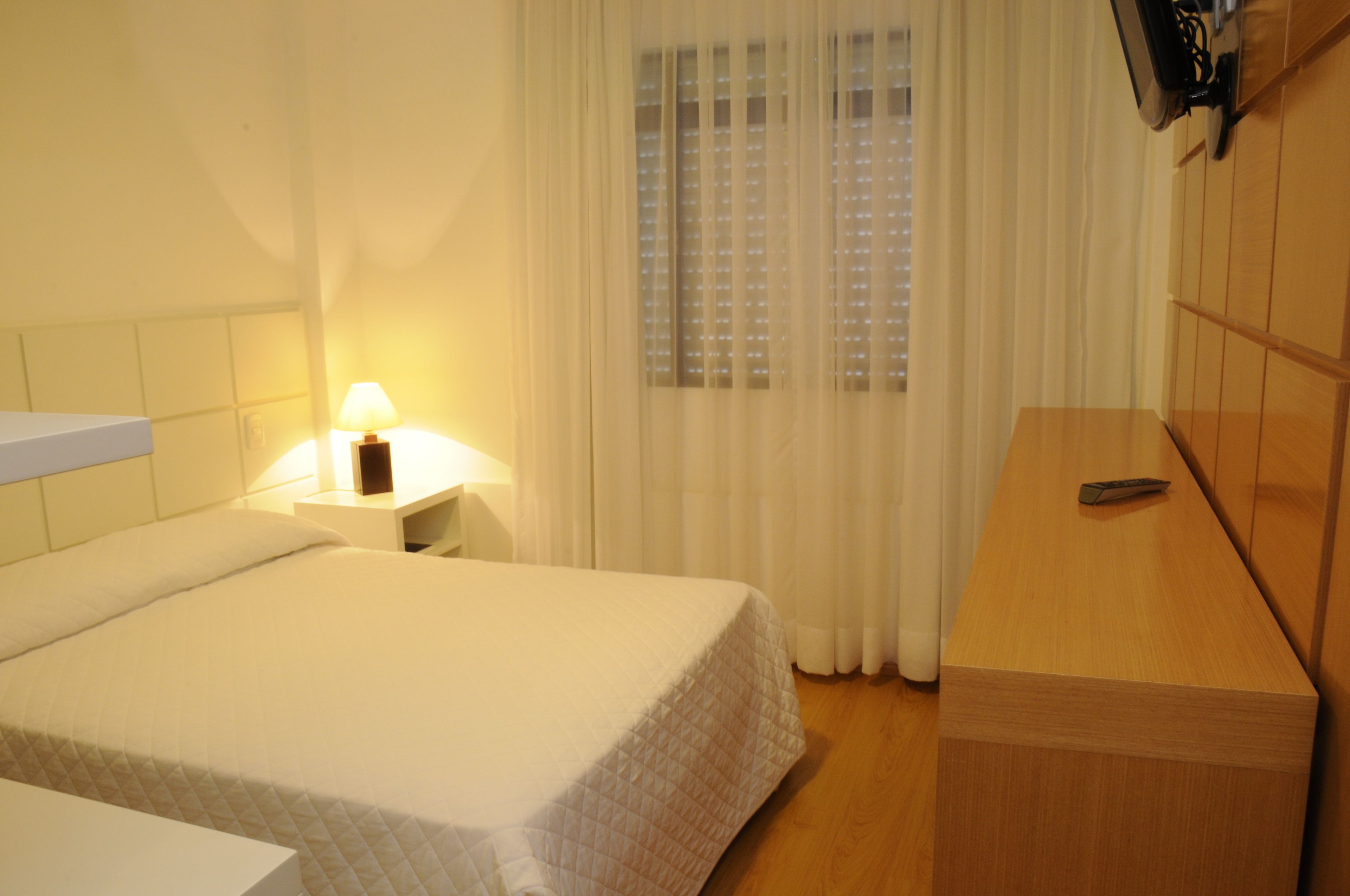 A hotel room with a bed, a bedside table with a lamp, a wooden dresser, and a window with blinds.