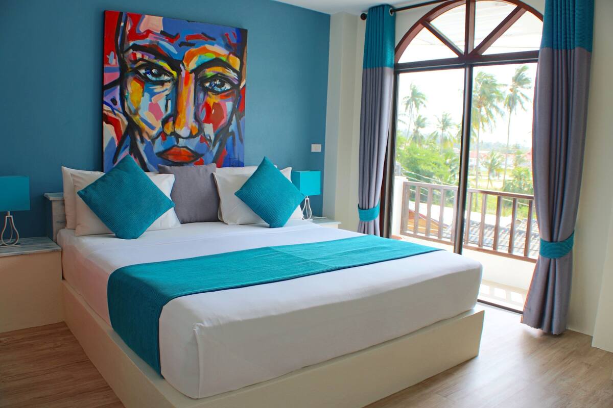 A modern hotel room with a large bed, a painting on the wall, and a balcony with a view of palm trees.