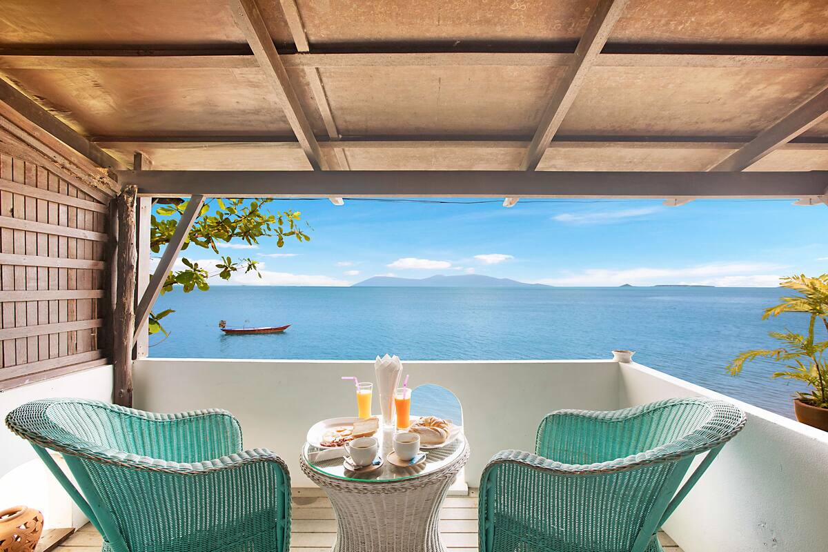 A balcony with wicker chairs, a table with breakfast items, and a view of the sea and mountains.