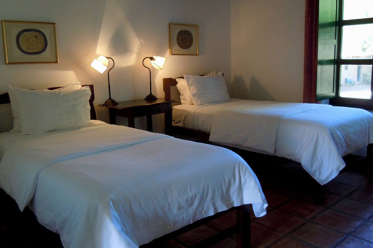 Deux lits simples avec du linge de lit blanc, une table de chevet avec une lampe et une fenêtre donnant sur l’extérieur.