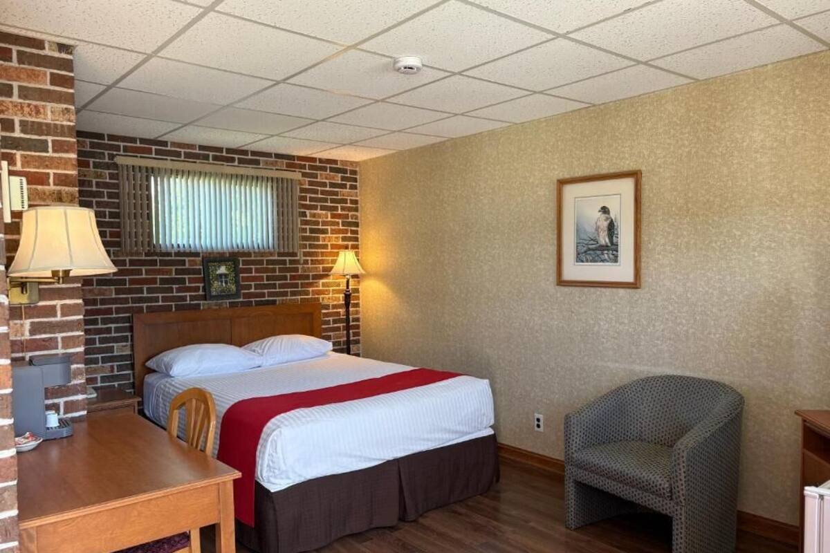 Hotellihuone, jossa on yksi seinä, joka on koristeltu tiilillä, valkoisilla lakanoiden peittämä sänky, puinen työpöytä tuolilla, kuviollinen nojatuoli ja seinällä oleva kehystetty kuva.