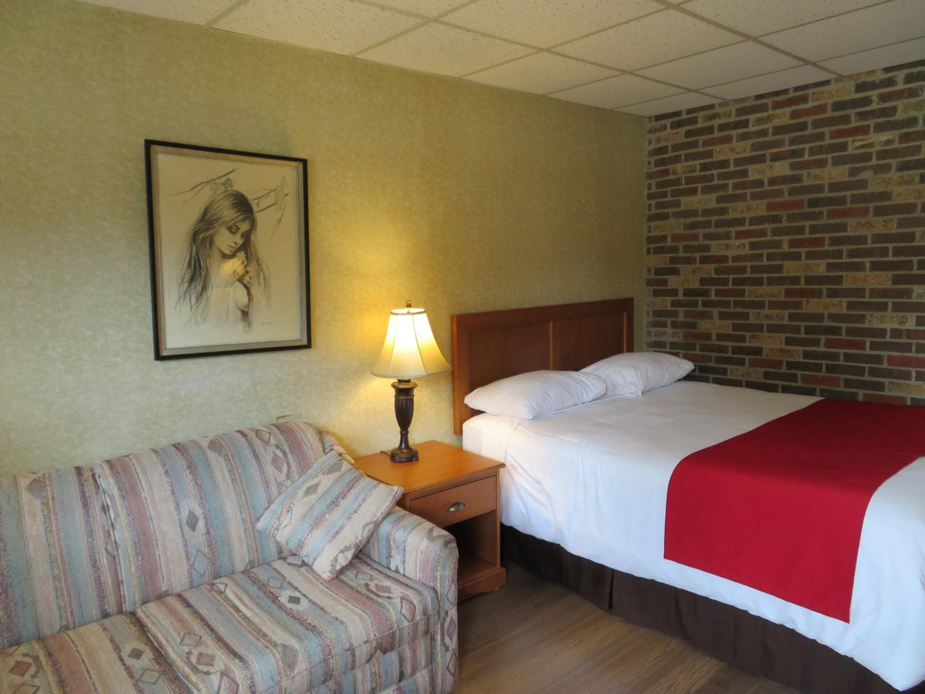 A hotel room with a bed, a nightstand with a lamp, a sofa, and a brick-patterned wall.