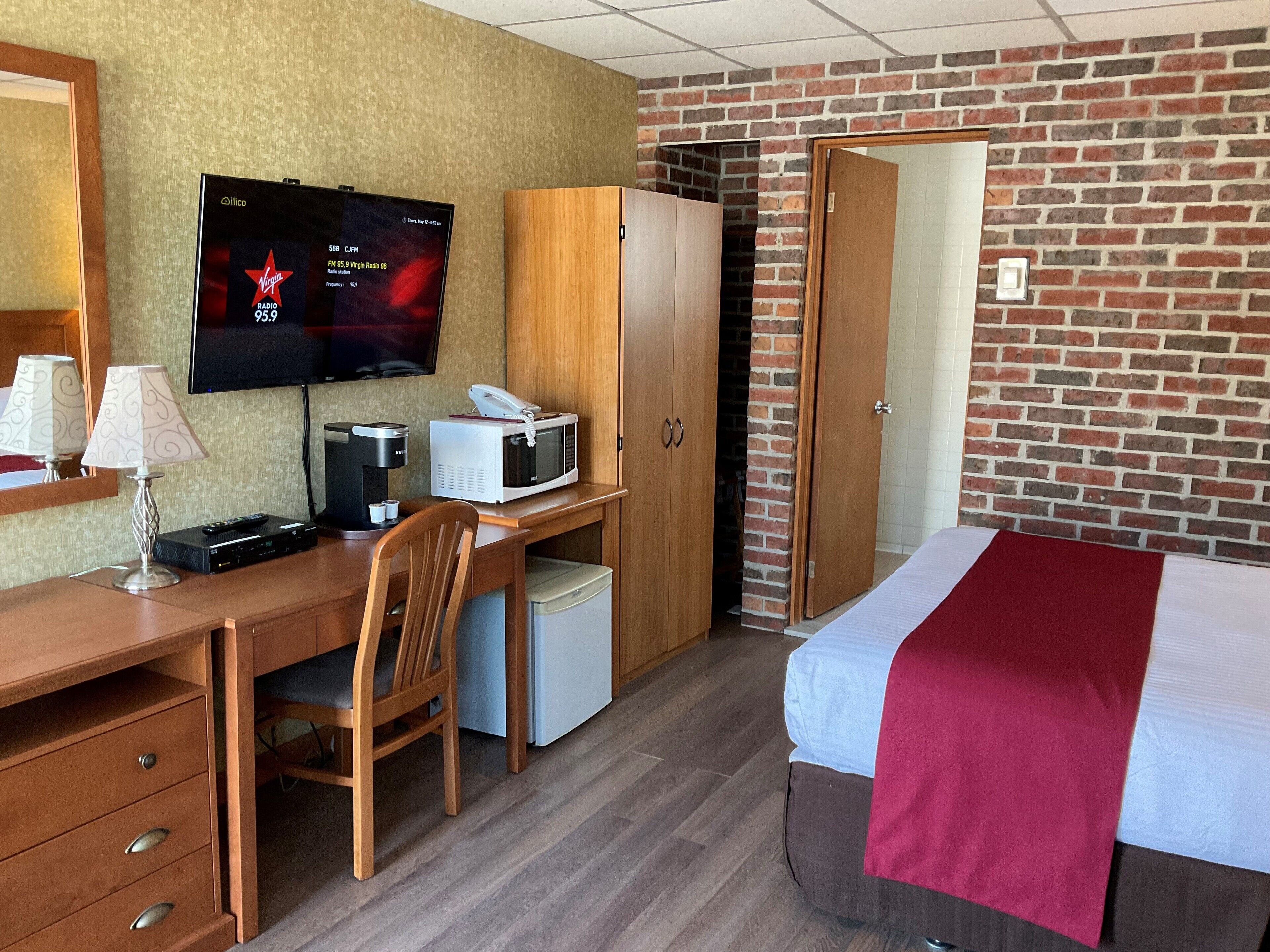 A hotel room with a wooden desk, a television, a microwave, a wardrobe, a bed, and a brick wall.