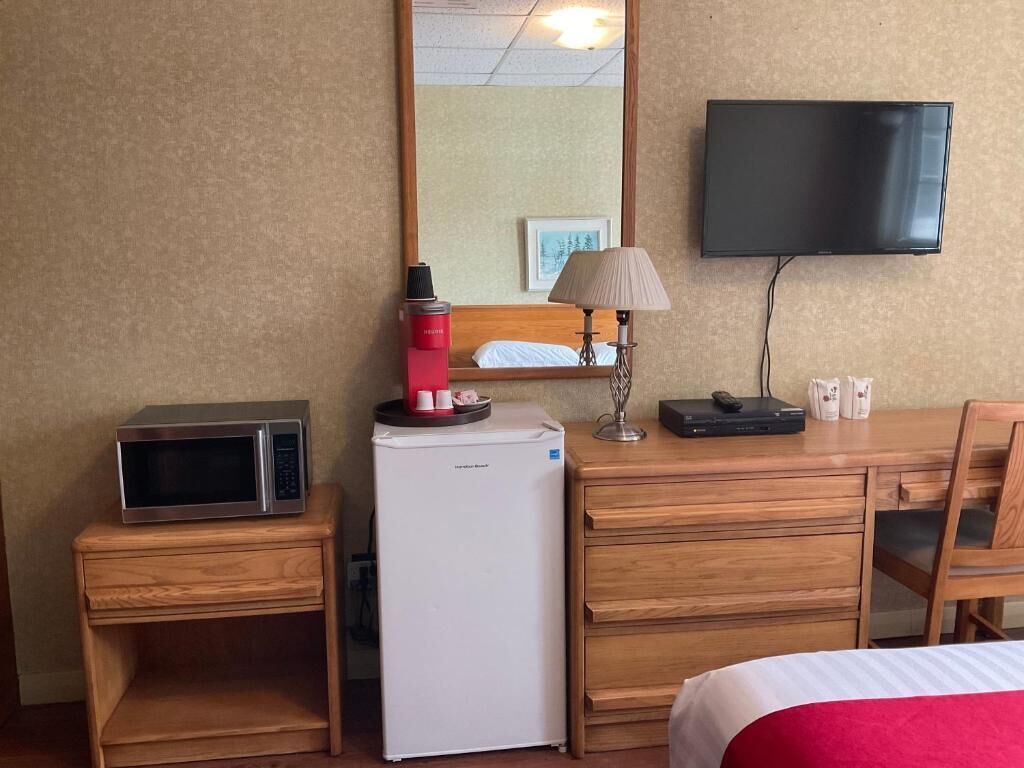Ein Hotelzimmer mit Bett, einem Nachttisch aus Holz mit Mikrowelle und kleinem Kühlschrank, einem Schreibtisch mit Lampe sowie einem an der Wand befestigten Flachbildfernseher.